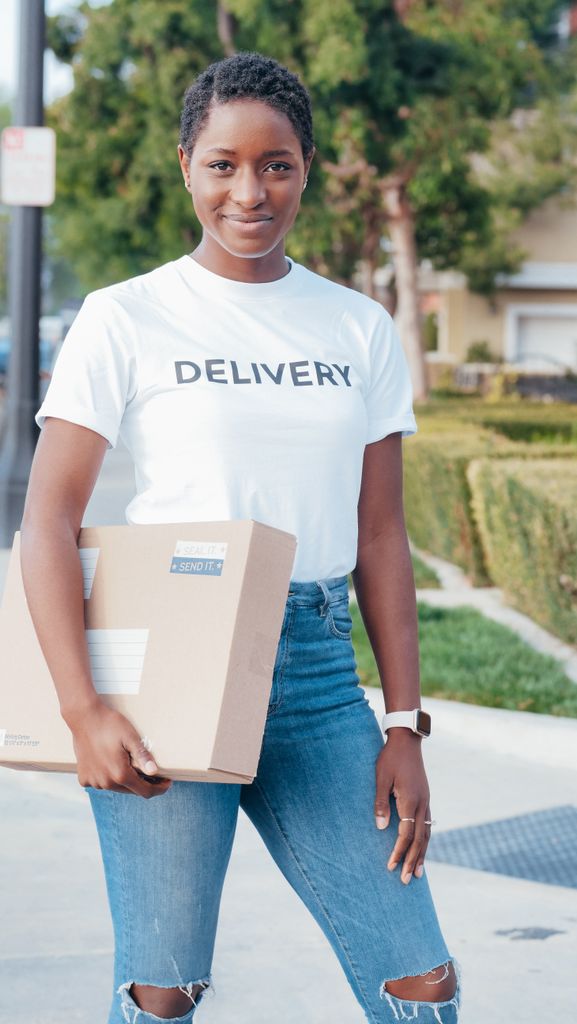 A young delivery woman smiles while holding a package outdoors, wearing a delivery t-shirt and jeans.